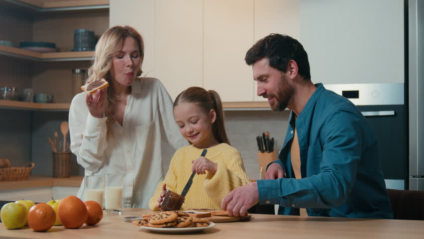 Happy Caucasian family dad father man help custody adopted child girl daughter smear peanut chocolate pasta on slice of bread mother mom eat delicious sandwich cooking breakfast together at kitchen