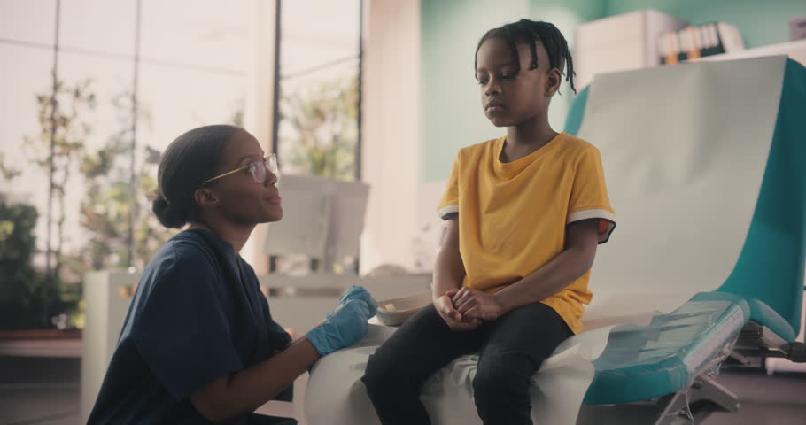 Young African American Boy Sitting In The Chair In Bright Hospital And Getting His Polio Vaccine. Female Black Nurse Is Performing Injection. Professional Woman Talks To Worried Kid And Calms Him Down
