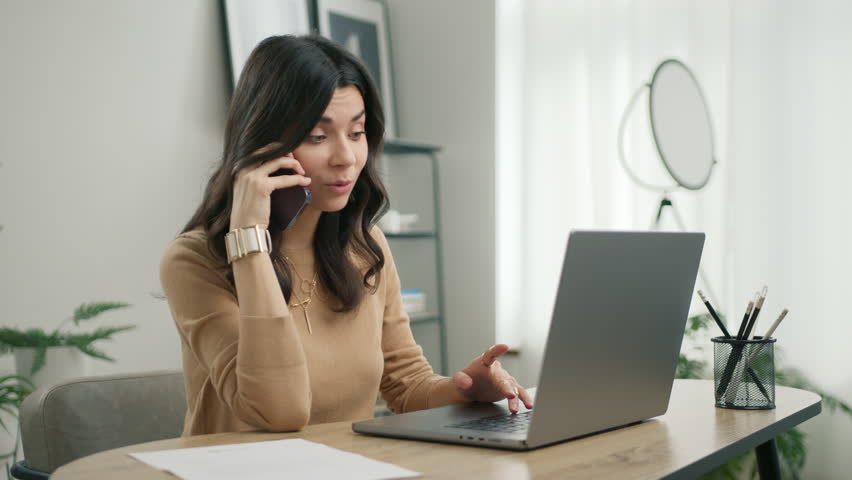 Positive latin female customer make mobile call confirming online website shopping order delivery concept. Smiling young hispanic business woman talking on phone using laptop sits at home office desk Royalty-Free Stock Footage #1104682937