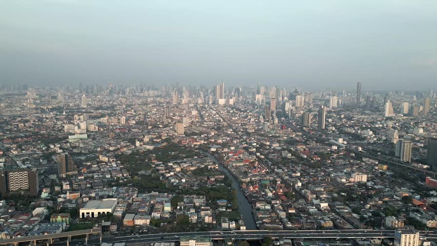 Stunning aerial view of the Bangkok city skyline with towering skyscrapers in the distance, and the scenic Chao Phraya River canal just before sunset. Royalty-Free Stock Footage #1104693355