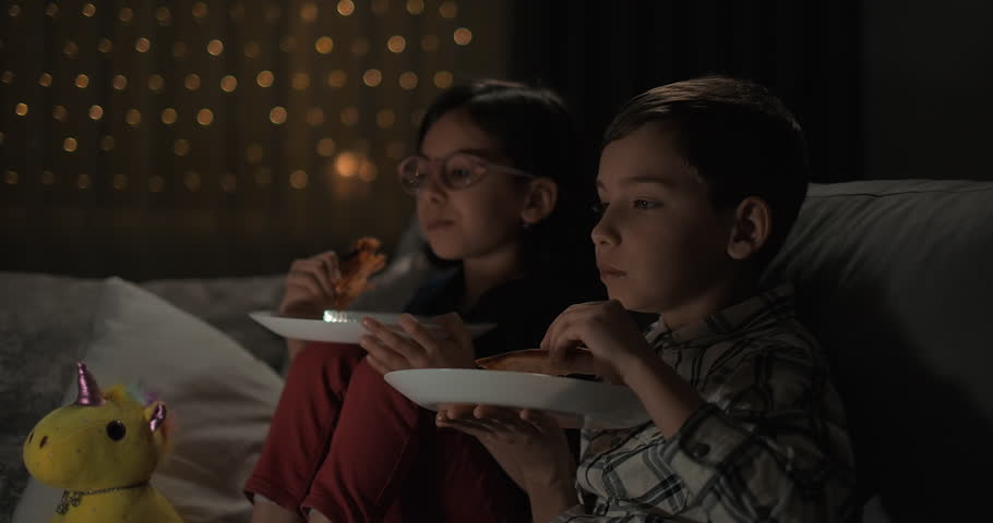 Kids watching TV eating pizza sitting on couch at home at night. Focused girl and boy watching movie cartoon at home | Shutterstock HD Video #1104702699
