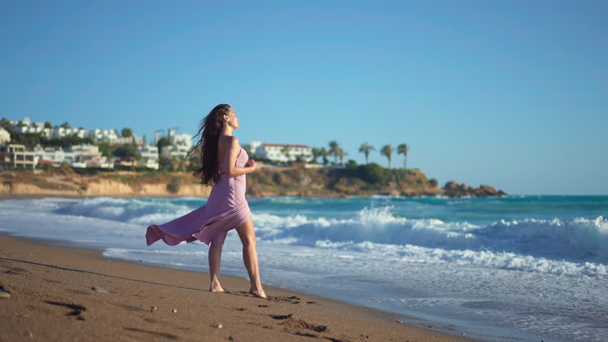 Wide shot young woman admiring azure blue Mediterranean sea waves rolling on beach in slow motion. Side view portrait of smiling happy Caucasian tourist in sunshine on ocean coast on Cyprus | Shutterstock HD Video #1104703657