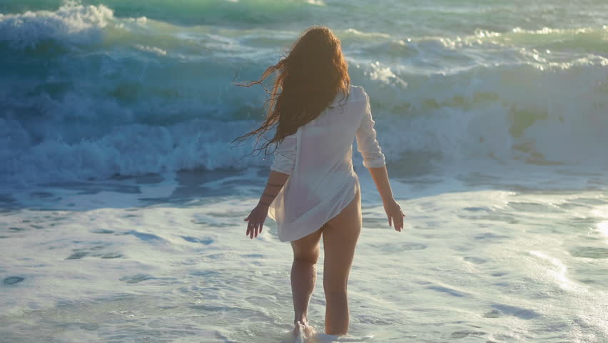 Young woman walking into waves of Mediterranean sea falling leaving in slow motion. Back view relaxed carefree female Caucasian tourist strolling in ocean at sunset