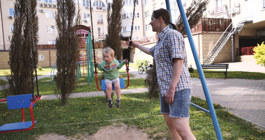 A two year old blonde toddler is riding on a swing. A young nanny is riding a baby on a swing in the playground. The baby is happy and having fun on the swing. | Shutterstock HD Video #1104705367