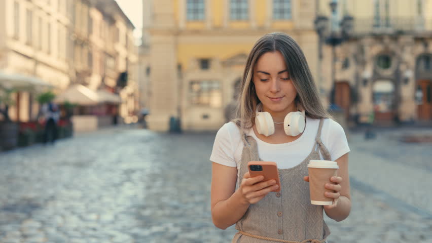 Attractive Caucasian Woman Reading Good News while Walking in the City Centre. Happy Tourist Girl Walking on the Street with Coffee and Modern Smartphone in Hands. Concept of Technology and Lifestyles | Shutterstock HD Video #1104706013
