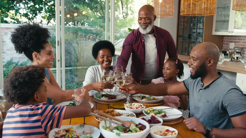 Multi-generation family sitting around table at home celebrating and doing cheers with glasses of wine and water before eating meal - shot in slow motion