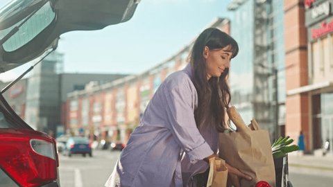 Girl carries products from cart to trunk. Woman has made purchases and is loading them into car. Young girl went shop to do grocery shopping. Girl with food in cart at background of infrastructure., videoclip de stoc