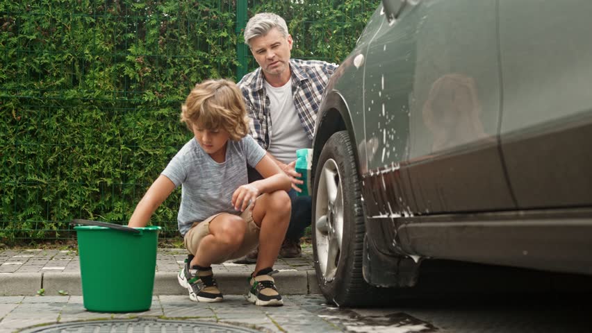 Little cute boy is sitting and washing car together with his dad at backyard. Gray haired man using sponges for teaching son to clean auto. Father is spending time with family members. Royalty-Free Stock Footage #1104711127