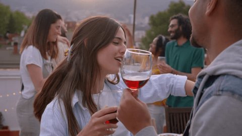 Joyful multi-ethnic couple dancing and drinking red wine celebrating rooftop party at sunset. Excited young friends having fun in background. People outdoors on summer vacation.  วิดีโอสต็อก
