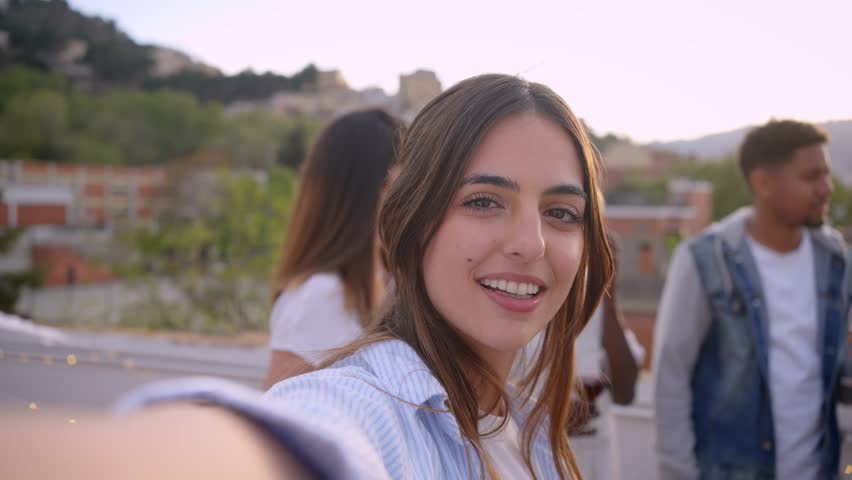 POV selfie of smiling young European girl looking camera shooting video for social media at rooftop party with friends. Groups of persons dancing and drink wine glasses. Relationships in youth people | Shutterstock HD Video #1104712689