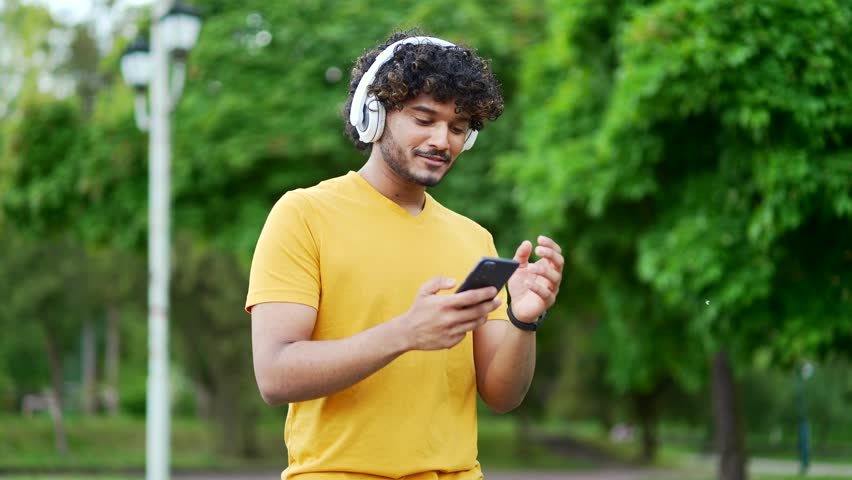 Young smiling student walking with headphones mobile phone enjoying listening to music outdoors on urban city park or trees backgroun. Happy male athlete in yellow t-shirt relaxing after training Royalty-Free Stock Footage #1104725633