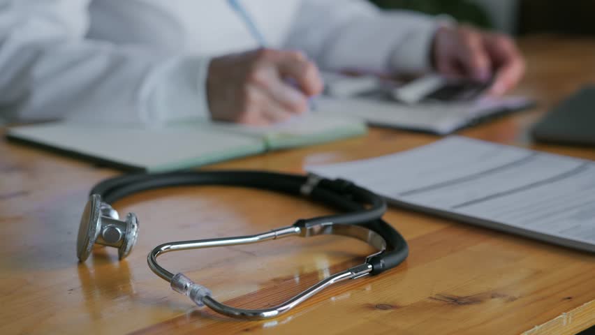 Close-up of medical equipment stethoscope with a doctor in the background. A professional doctor in a white coat is working at a table. The concept of general practice. High quality 4k footage Royalty-Free Stock Footage #1104726961