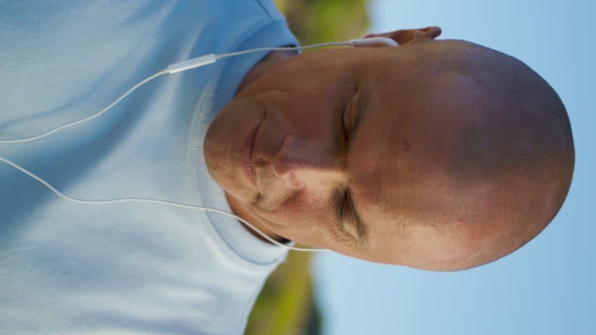 Happy man listening earphones music closeup. Relaxed athlete enjoying mountains vacation using headphones at beautiful landscape. Fit guy resting after workout vertical view. Active morning lifestyle.