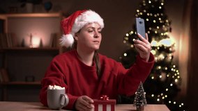 Young woman opening christmas present and feeling disappointed while having video call with friends or family. Girl sitting near christmas tree unpacking gift and looking unhappy