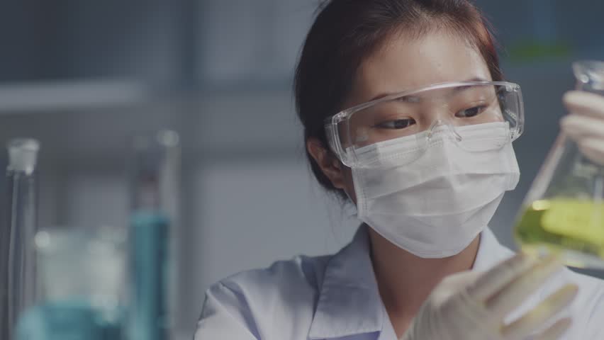 Scientist wearing white gloves and safety glasses is writing analysis of a chemical sample in the scientific research laboratory. The scientist holding an erlenmeyer flask containing yellow liquid Royalty-Free Stock Footage #1104733803
