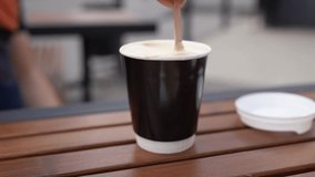 Slow and fast stirring sugar in a glass in a street cafe. Hand holding a wooden spoon and quickly stir cappuccino with white milk foam in a black paper cup outdoor. Video footage coffee to go concept