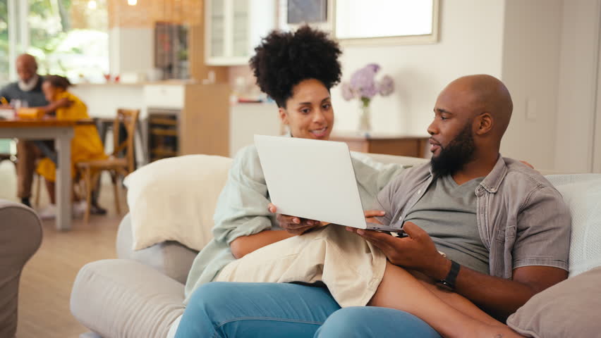 Couple at home looking at laptop together booking holiday tickets, checking finances or shopping online with multi-generation family in background - shot in slow motion Royalty-Free Stock Footage #1104742777