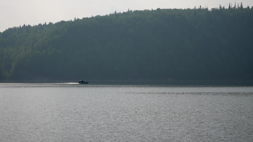 Remote view from coast of speedboat sailing across lake on background of rolling hills of green trees on cloudy day. Tracking shot of high-speed motorboat floating wide river. Shooting in slow motion.
