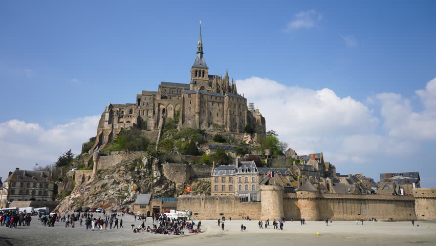 Tourists walking around by Le Mont Saint-Michel castle on sand of low tide on sunny day on background of blue sky. Crowd of people strolling on sand sea, seagulls flying though air, slow motion.