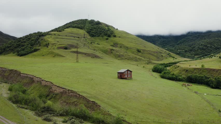 A lonely wooden house on stilts in a field in a mountain valley among green hills. Beautiful view of the highlands from a quadcopter