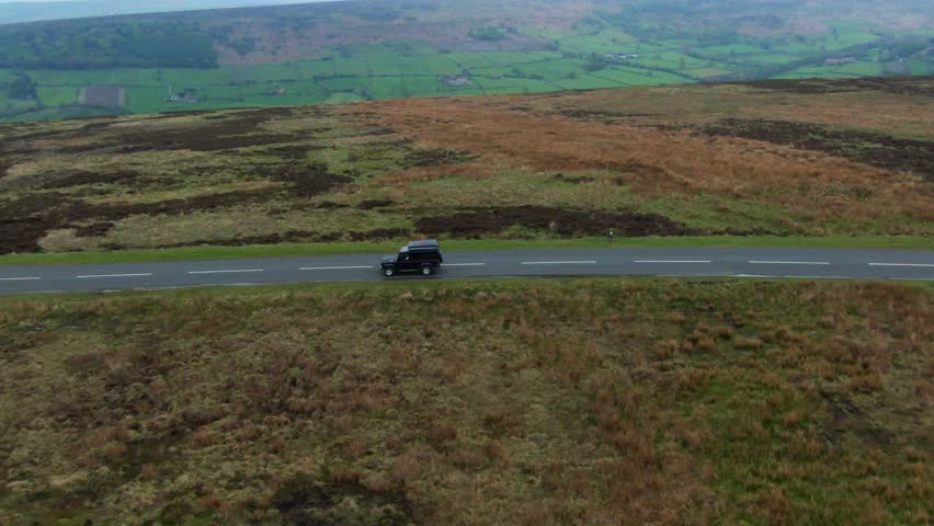 Black off road 4x4 truck driving on a empty country road along the top of a mountainous hill range overlooking at green valley on overcast cloudy day to reveal a long winding empty road for driving