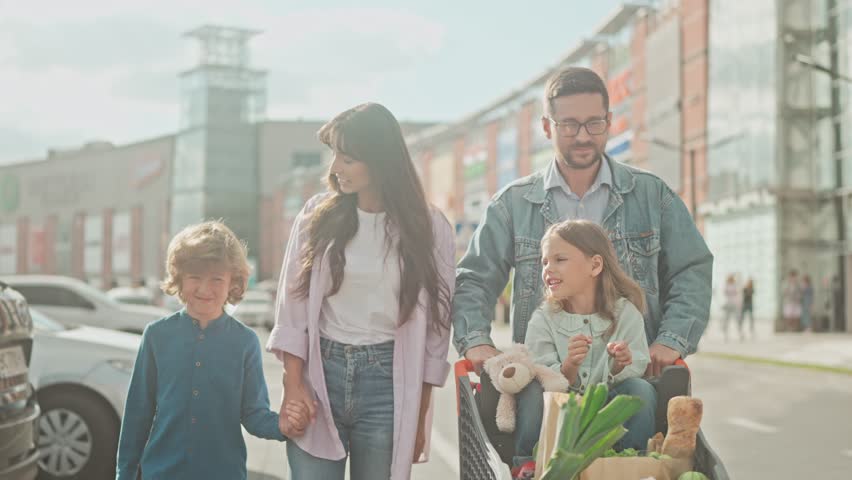 Happy family made groceries at food market. Mom keep son by hand and go to car parking. Dad push daughter in grocery cart. Girl is pointing finger at something to dad. Leisure of children and parents. Royalty-Free Stock Footage #1104753221