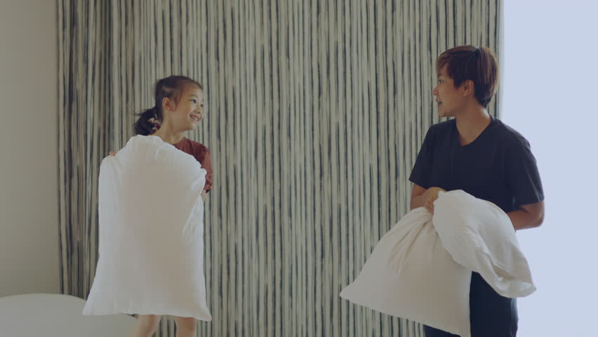 Slow motion 4K of adorable asian daughter and mom who are cheerfully playing pillow battle on the bed with emotional expression of happiness, joyfulness and fun shows concept of parental freedom.