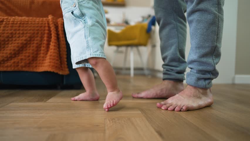 Baby first steps.Toddler at home takes funny first steps.Dad teaches child to walk, legs close-up.Toddler feet on laminate floor.Father's day child education.Kid dream study at home.walk baby learning Royalty-Free Stock Footage #1104756645