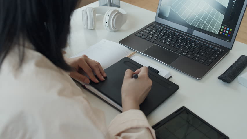 Over-shoulder arc shot of unrecognizable Asian female engineer sitting at desk, working with stylus on graphic tablet, and manipulating solar panel model on laptop screen Royalty-Free Stock Footage #1104761103