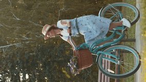 Cheerful female in dress and straw hat riding bicycle in golden sunset lights. Vertical video. Young charming woman cycling on retro vintage bicycle in green city park at summer sunset