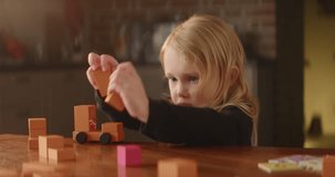Busy game on the table performed by cute baby girl, orange wooden toy blocks, shy autistic child plays alone, builds street with houses. Mental disorder, domestic background. High quality 4K footage