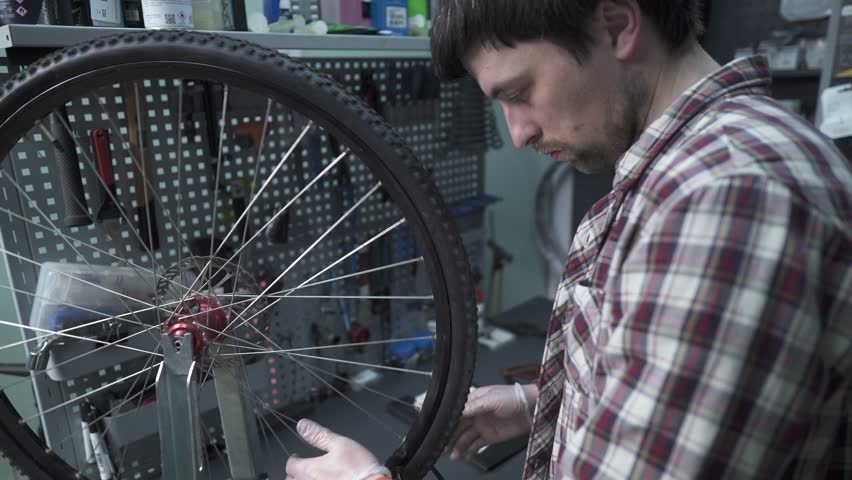 A male bicycle mechanic aligns the tension spokes of a bicycle wheel with a special tool at the workplace in the workshop of a bicycle shop. Bicycle maintenance and service. Wheel rim alignment tool. Royalty-Free Stock Footage #1104776083
