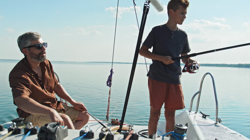 Mature father watching his teen son fishing in lake using rod during trip on sailboat on sunny summer day Royalty-Free Stock Footage #1104777477