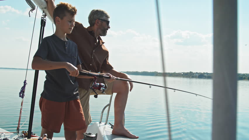 Mature Caucasian man wearing sunglasses watching his teen son fishing in lake using spinning rod during trip on sailing yacht Royalty-Free Stock Footage #1104781523