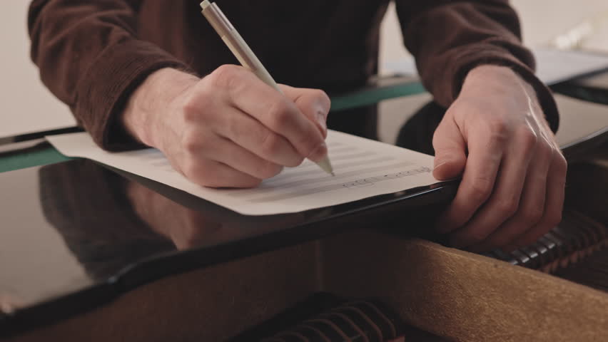 Slowmo closeup of unrecognizable male songwriter writing musical notes for piano on sheet of paper while creating song sitting alone indoors
