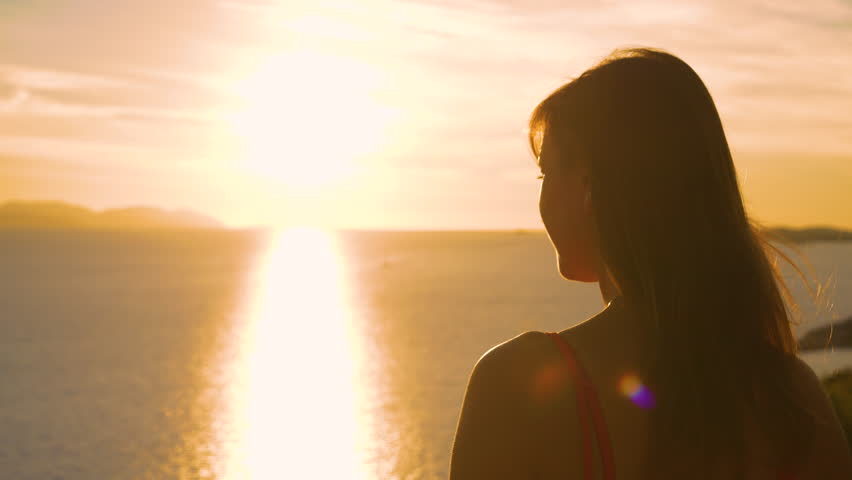 LENS FLARE, CLOSE UP: Pretty lady stretching her arms while watching sunrise. She is admiring stunning view and watches first rays of summer sun gently spilling over picturesque Dalmatian seascape. Royalty-Free Stock Footage #1104783765