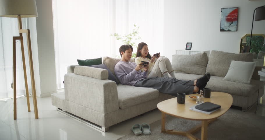 South Korean Couple Sitting on a Cozy Couch, Playing Funny Arcade Video Games Together in a Stylish Bright Living Room. Beautiful Couple in a Relationship Enjoying Time on a Sofa at Home Royalty-Free Stock Footage #1104788177