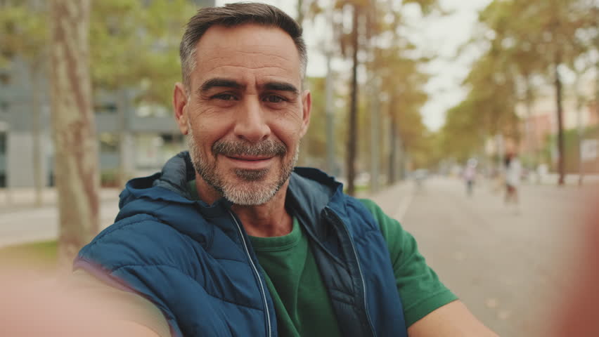 Close-up, mature man wearing casual clothes, sitting on bench in the park, talking on video call from smartphone, waving his hand in greeting