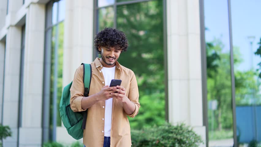 Handsome smiling student with a backpack uses a smartphone while walking in the campus space near university building. Male chats online with friend, browses social networks, writes or reads messages Royalty-Free Stock Footage #1104794583