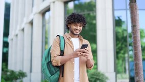 Handsome smiling student with a backpack uses a smartphone while walking in the campus space near university building. Male chats online with friend, browses social networks, writes or reads messages - Powered by Shutterstock - Get 15% off with code: PIKWIZARD15