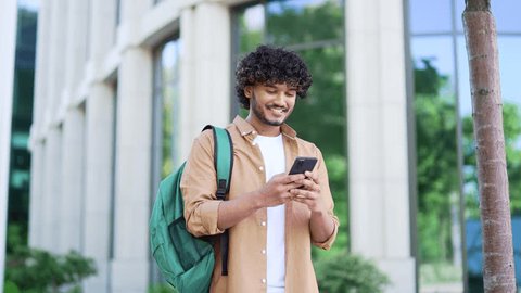 Handsome smiling student with a backpack uses a smartphone while walking in the campus space near university building. Male chats online with friend, browses social networks, writes or reads messages 库存视频
