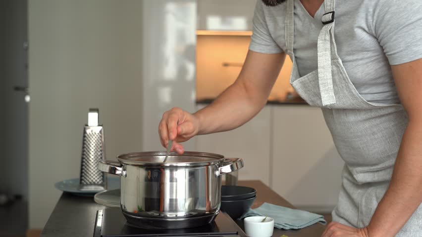 Man stirs boiling soup with spoon in pot on stove and tastes on background of home kitchen. Cooking food