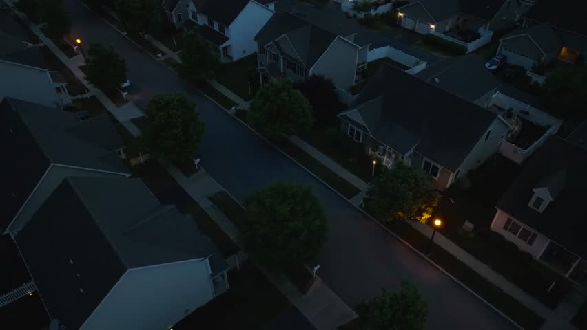 American houses and homes at night with street light and trees. Aerial orbit and rotational tilt up reveal of large neighborhood in summer during blue hour dusk.
