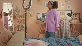 Medium shot of 10 year old African American cute girl recording trendy dancing video on smartphone in her bedroom using ring light and tripod