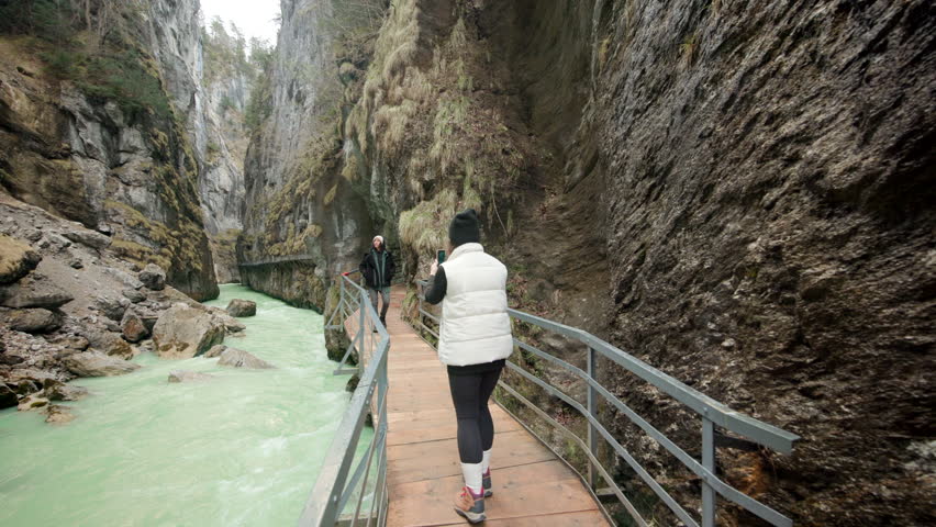 A blonde girl in hat and warm white vest takes a picture on her phone of a lady in jacket walking towards her on the wooden bridge, embraced by the majestic mountains of Aare Gorge, Switzerland.