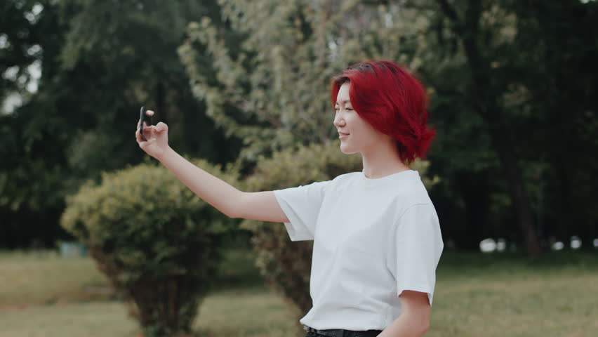 Young asian girl using a smartphone to make a video for her friends on social media while she is walking outdoors in the city park. Concept of stylish youth who use gadgets