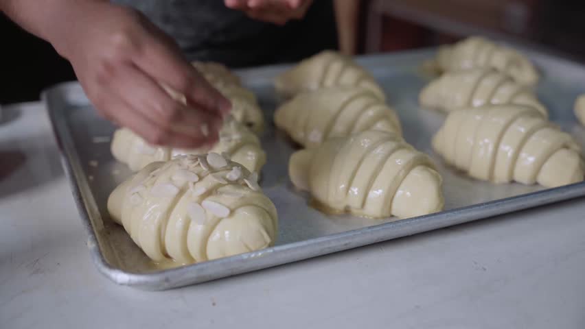 A young Hispanic baker is covering croissants with almond toppings before baking