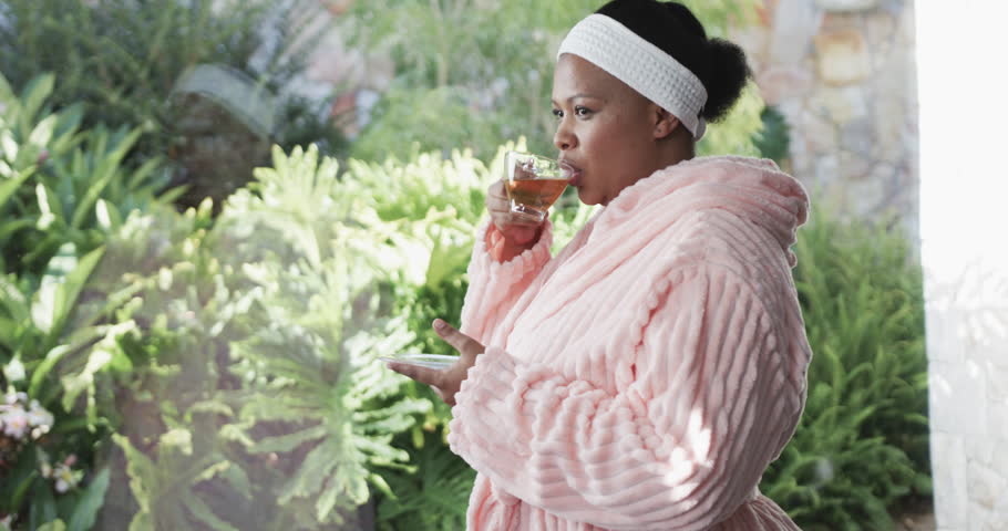 African american plus size woman wearing bathrobe and drinking tea at spa, unaltered, slow motion. Spa, beauty, relaxation and body inclusivity. Royalty-Free Stock Footage #1104813559