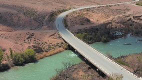 Picturesque mountain aerial view of bridge crossing scenic river reflective blue sky. Kyrgyzstan. Central Asia. Video of drone views of river crossing bridge. 