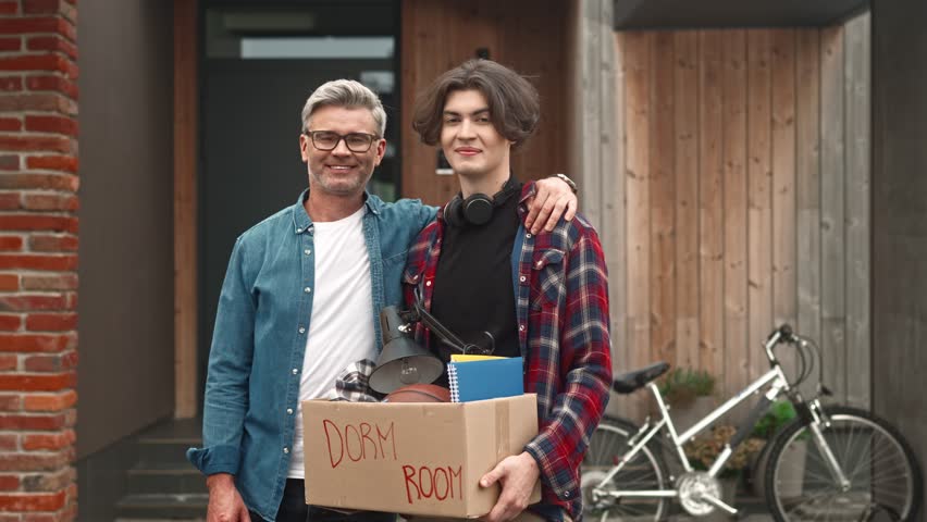 Portrait of adult man and young boy standing outside and looking at camera with smile. Dad hugs his son leaving parents' house. Cute guy entering university and moving away from home.